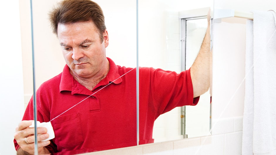 man standing in the bathroom in front of a mirror pulling out a long strand of dental floss
