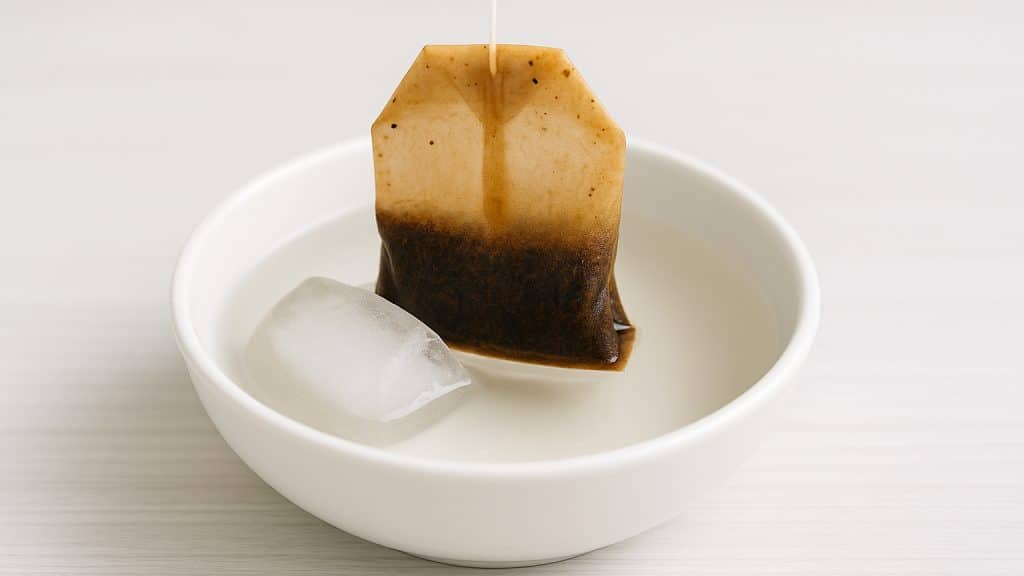 Black tea bag soaking with an ice cube in a small bowl, used as a home remedy after a tooth extraction