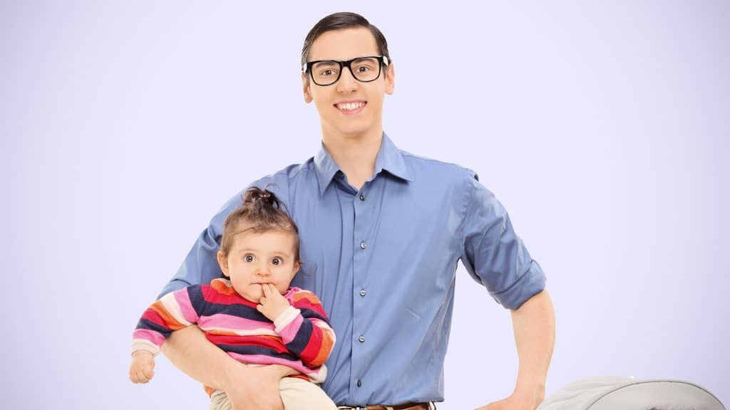 Smiling man in glasses holding a baby with fingers in her mouth, standing against a soft lavender background.